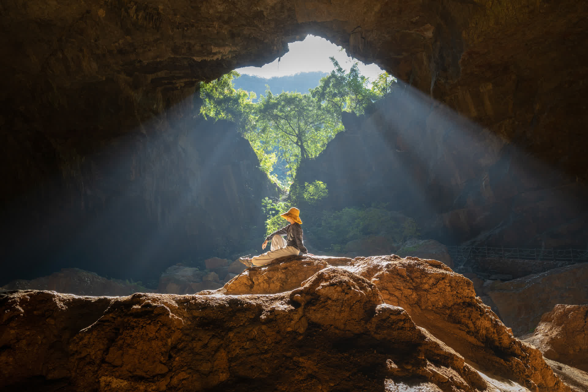 A traveler in a sun hat resting on a rock inside a Mexican cenote as light rays pour through the cave mouth.